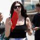 A woman on the street waves a red fan and holds her phone. She wears sunglasses and a black top.