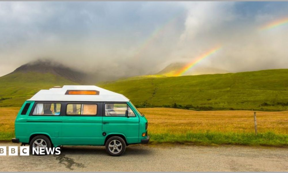 A traditional-style VW campervan parked on a road next to fields and mountains. They are slightly obscured by low cloud, but a rainbow is also visible in the background.