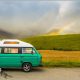 A traditional-style VW campervan parked on a road next to fields and mountains. They are slightly obscured by low cloud, but a rainbow is also visible in the background.