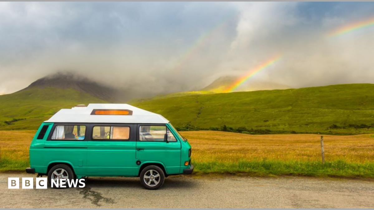 A traditional-style VW campervan parked on a road next to fields and mountains. They are slightly obscured by low cloud, but a rainbow is also visible in the background.