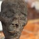 An Egyptian mummy head at the forefront of a blurred background. The skin looks rough, dry and black.  Its eyes are closed.  You can see a few yellow teeth through the gap between its lips.