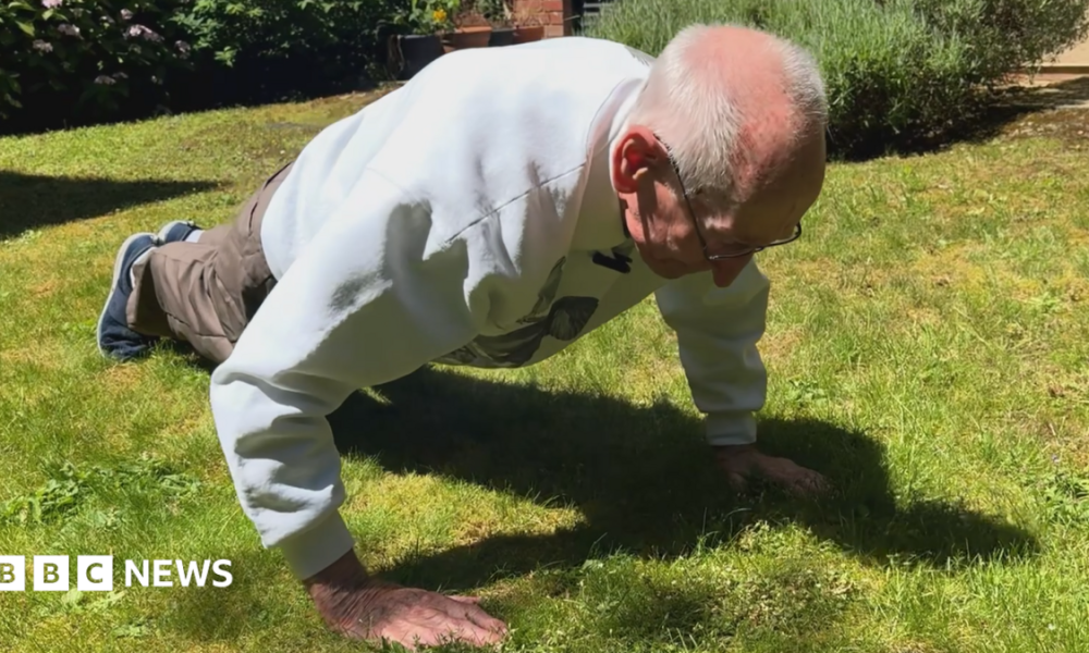 An elderly man doing a press-up on grass in a garden.