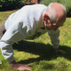 An elderly man doing a press-up on grass in a garden.