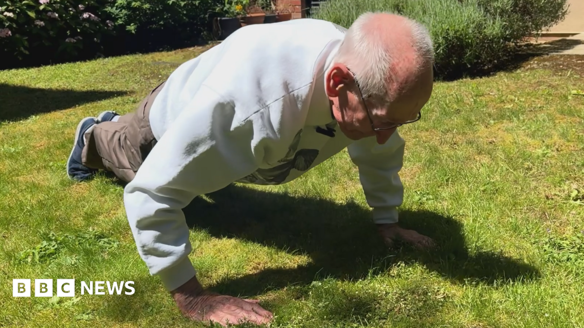 An elderly man doing a press-up on grass in a garden.