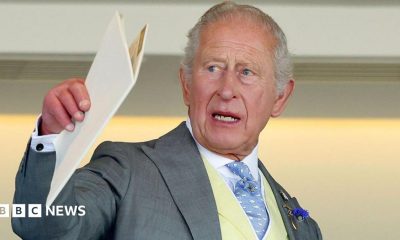 King Charles holding a white A4 booklet and gesturing with it. He is wearing a pale great suit, cream waistcoat, blue and white patterned tie, and a white shirt.