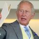 King Charles holding a white A4 booklet and gesturing with it. He is wearing a pale great suit, cream waistcoat, blue and white patterned tie, and a white shirt.