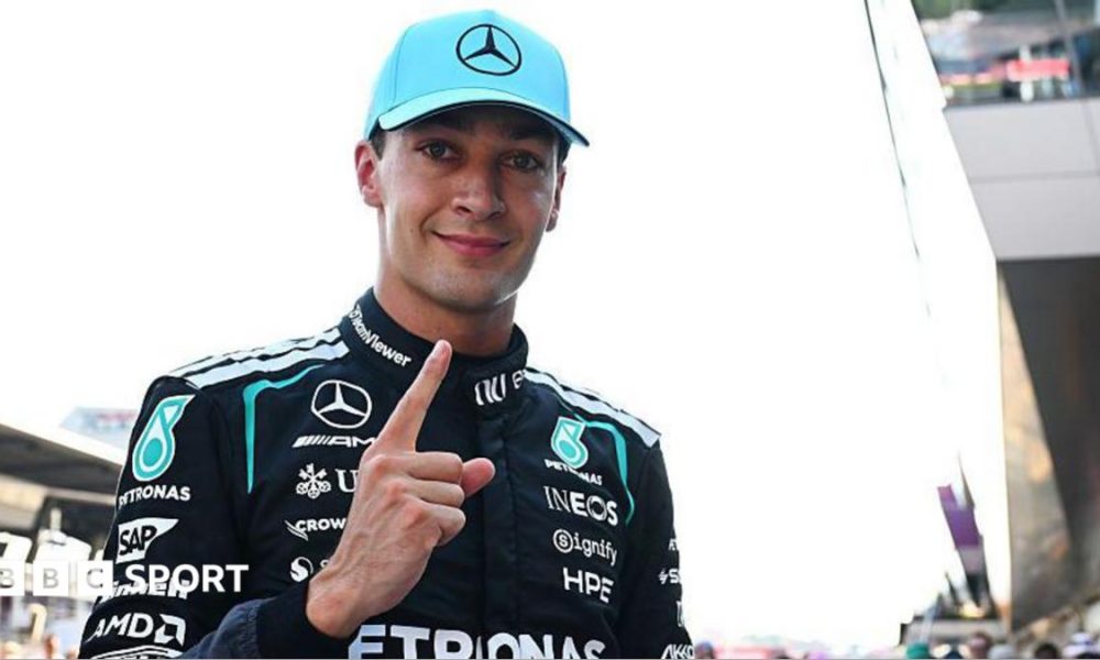 George Russell looks at the camera and points his index figure upwards