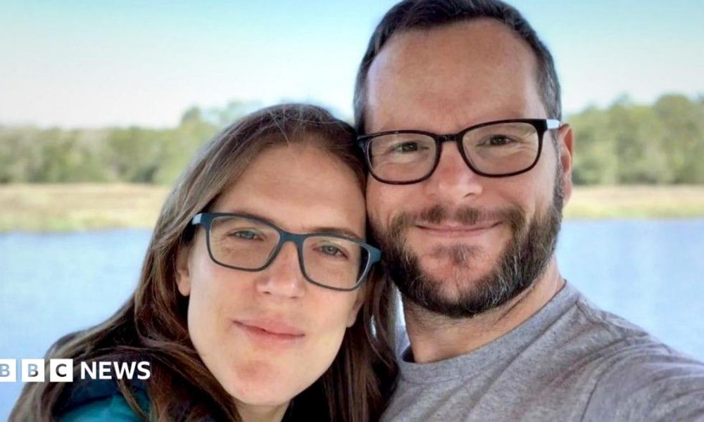 Katie and Alan Donegan smile at the camera while both wearing glasses during a selfie in front of a lake and trees against a blue sky.
