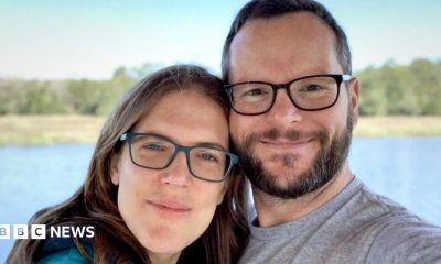 Katie and Alan Donegan smile at the camera while both wearing glasses during a selfie in front of a lake and trees against a blue sky.