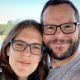 Katie and Alan Donegan smile at the camera while both wearing glasses during a selfie in front of a lake and trees against a blue sky.