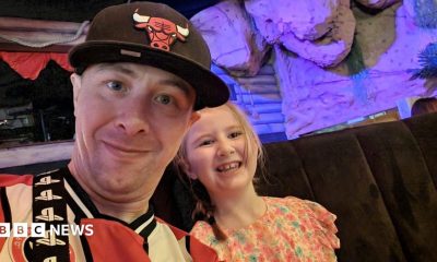 A man in a red and white football shirt and baseball cap smiles next to a younger girl in a pretty flowery dress
