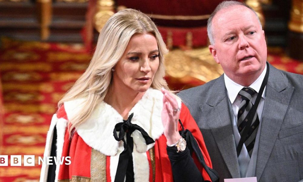A woman with long blonde hair is dressed in a fur trimmed red and gold gown and her middle aged husband is in a grey three-piece suit. They are standing inside a regal looking room with red and gold carpets