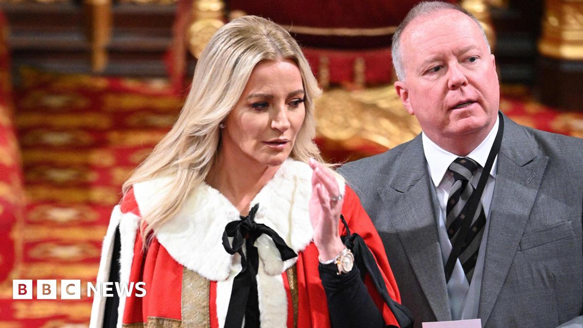 A woman with long blonde hair is dressed in a fur trimmed red and gold gown and her middle aged husband is in a grey three-piece suit. They are standing inside a regal looking room with red and gold carpets