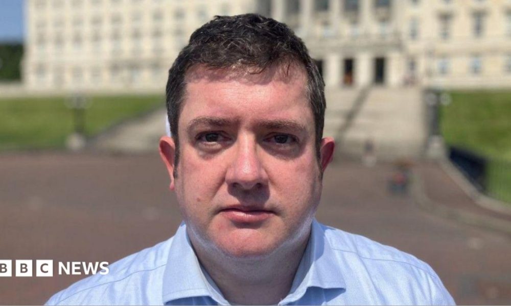 Dr David Farren, a man with dark hair, wearing a blue shirt, standing in front of Stormont Parlaiment Buildings.