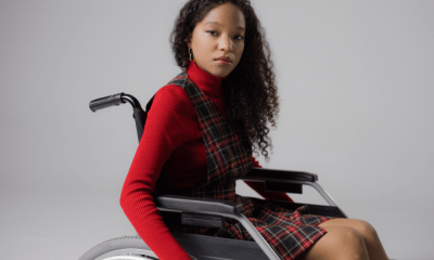 A disabled teenage girl, wearing lipstick to match her autumnal high neck top and tartan dress, looks at the camera with a neutral expression from her wheelchair in a studio