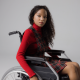 A disabled teenage girl, wearing lipstick to match her autumnal high neck top and tartan dress, looks at the camera with a neutral expression from her wheelchair in a studio