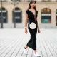Amanda Derhy wears earrings, a black low-neck v-neck dress, a white circular round straw summer bag, a watch, white Adidas sneakers shoes, on August 09, 2020 in Paris, France.