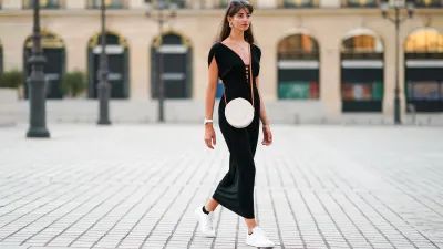 Amanda Derhy wears earrings, a black low-neck v-neck dress, a white circular round straw summer bag, a watch, white Adidas sneakers shoes, on August 09, 2020 in Paris, France.