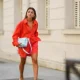 Alexandra Pereira wears earrings, a bold neon orange / red oversized shirt, matching mini shorts, a pale pastel blue woven leather bag from Bottega Veneta, pale pastel quilted leather sandals from Bottega Veneta, on July 20, 2021 in Paris, France.