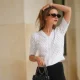 Segolene Hyppolite wears sunglasses, a white wool cardigan / top from Agnes B, a white flower shaped bracelet, a Chanel bag with floral texture / details, black pants from Agnes B, during a street style fashion photo session, on May 12, 2024 in Paris, France.