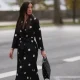A Fashion Week guest is seen wearing round diamond stud earrings; a long-sleeved black midi dress with white polka dot pattern, notched lapels, and a tie belt; a gold bangle; a rose gold and black watch from Rolex; a gold ring; and a rectangular black handbag with a white polka dot pattern before the Elie Saab Womenswear Spring/Summer 2025 Show as part of Paris Fashion Week on September 28, 2024 in Paris, France.