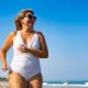Beautiful middle-aged woman in swimsuit running on beach against background of blue sky in summertime. Front view