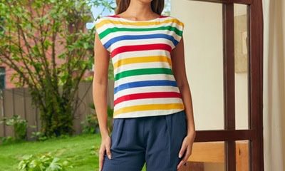A woman wearing a multicolored striped tee and navy shorts. She is barefoot and standing in front of a partially opened door in front of a yard.