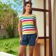 A woman wearing a multicolored striped tee and navy shorts. She is barefoot and standing in front of a partially opened door in front of a yard.