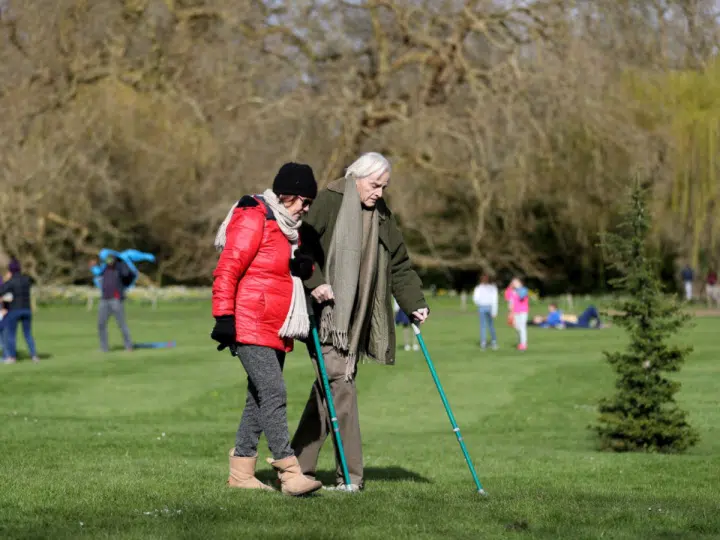 Carers Week: An older pair, one with two crutches, and the other locking their arm, walk and enjoy the spring sunshine at Mottisfont one of the National Trust parklands on 21 March 2020 in Romsey, UK