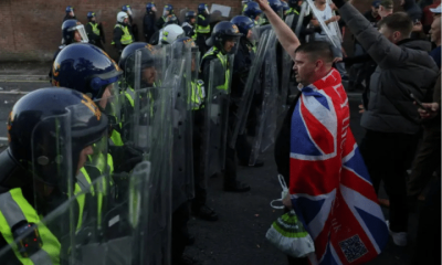 Armed riot police hold their shields up in a row while men, one at the front with Union Jack flag draped on his back, show Nazi salutes. Skwawkbox argues Israel-bought politicians show hypocrisy when responding to riots and protests