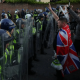 Armed riot police hold their shields up in a row while men, one at the front with Union Jack flag draped on his back, show Nazi salutes. Skwawkbox argues Israel-bought politicians show hypocrisy when responding to riots and protests