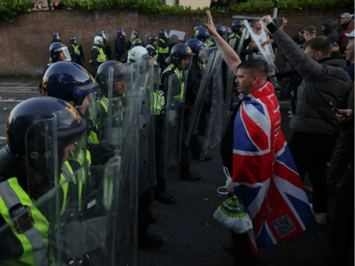Armed riot police hold their shields up in a row while men, one at the front with Union Jack flag draped on his back, show Nazi salutes. Skwawkbox argues Israel-bought politicians show hypocrisy when responding to riots and protests