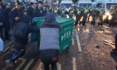 A still from a video posted on Young Bob's X account of two males pushing a large wheelie bin with a fire inside towards a row of armed riot police at far-right riots this week. Others line the pavements filming with their mobile phones and cheering the pair on