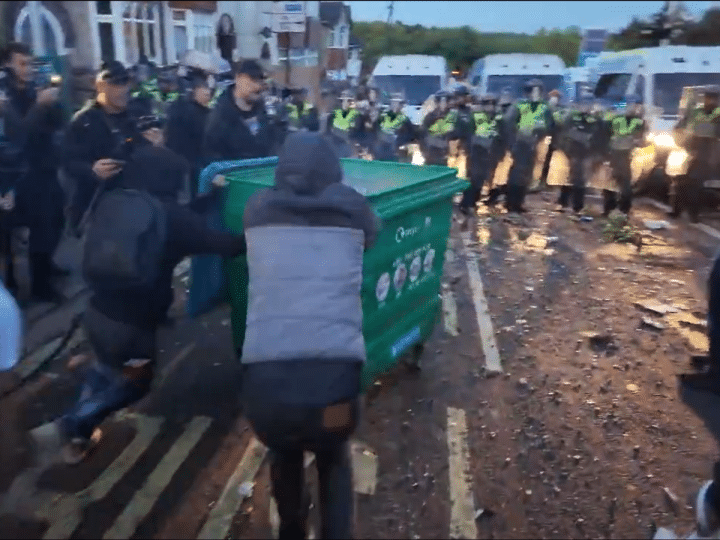 A still from a video posted on Young Bob's X account of two males pushing a large wheelie bin with a fire inside towards a row of armed riot police at far-right riots this week. Others line the pavements filming with their mobile phones and cheering the pair on