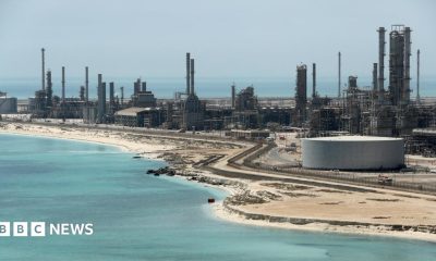 A file photo of Aramco's Ras Tanura oil refinery and oil terminal, which lies along a stretch of coast. The site comprises a network of several tall chimney-shaped structures and smaller buildings. It is daytime and the water along the coastline is a clear blue.