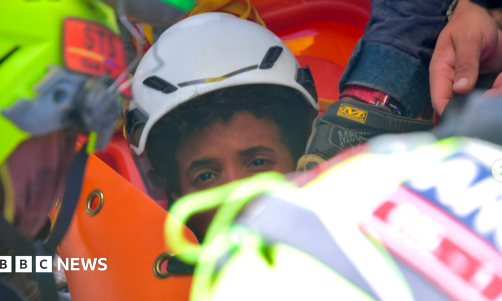 the picture shows half of the boys face - the rest is obscured by rescue workers. He's wearing a white hard hat and has dark curly hair.