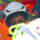 the picture shows half of the boys face - the rest is obscured by rescue workers. He's wearing a white hard hat and has dark curly hair.