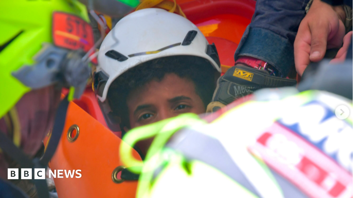 the picture shows half of the boys face - the rest is obscured by rescue workers. He's wearing a white hard hat and has dark curly hair.