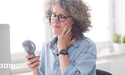 A woman sits at her desk looking uncomfortable, while using a handheld fan to cool down