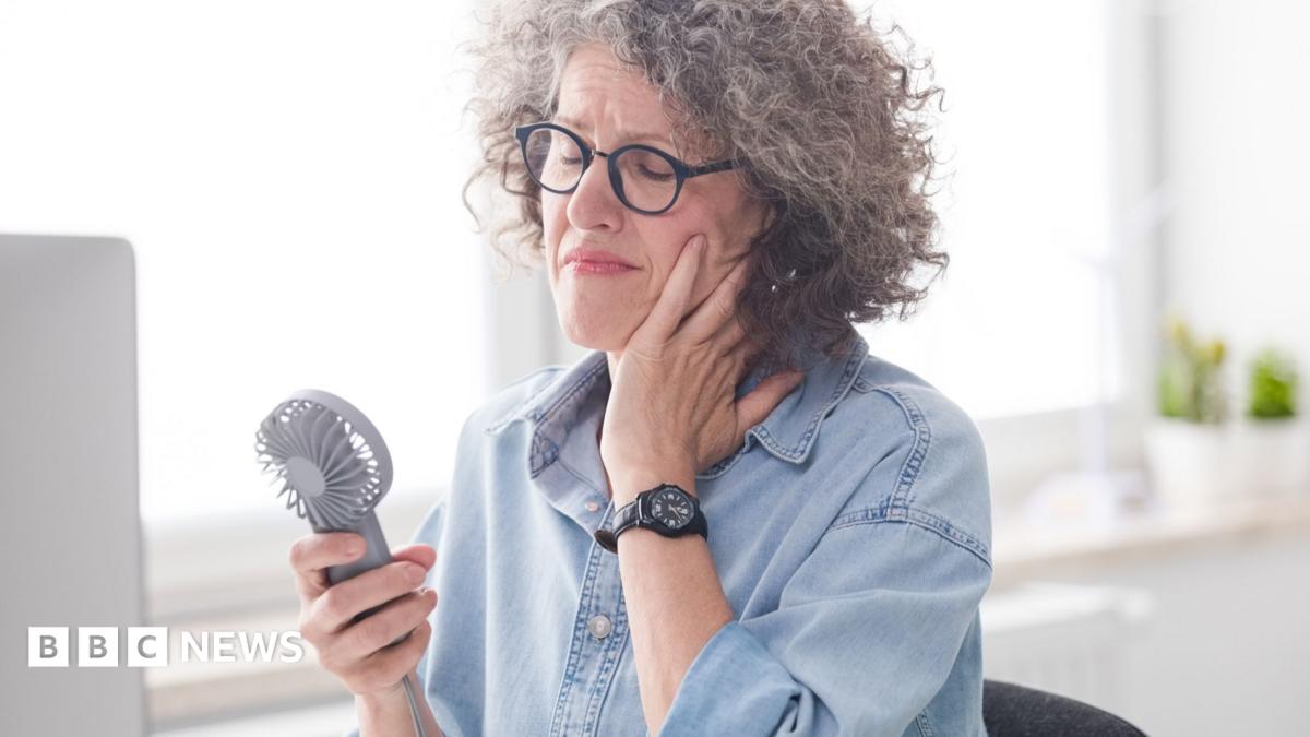 A woman sits at her desk looking uncomfortable, while using a handheld fan to cool down