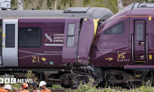 Two damaged trains, the front of one purple train is crushed into the end of another, with severe impact damage visible around the cab area.
