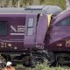 Two damaged trains, the front of one purple train is crushed into the end of another, with severe impact damage visible around the cab area.