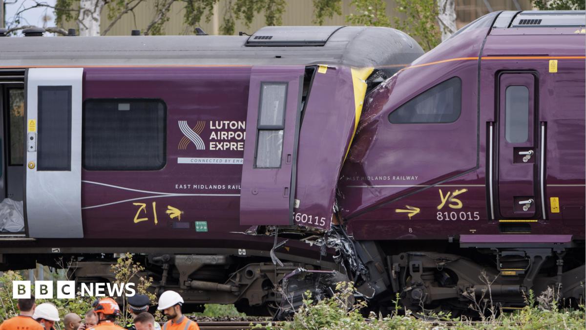 Two damaged trains, the front of one purple train is crushed into the end of another, with severe impact damage visible around the cab area.