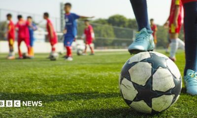 A close up of a football with a young player's foot on it. Other children are in the background standing on the fenced green play area.