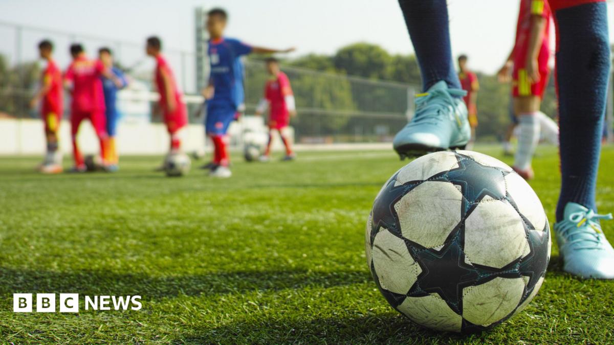 A close up of a football with a young player's foot on it. Other children are in the background standing on the fenced green play area.