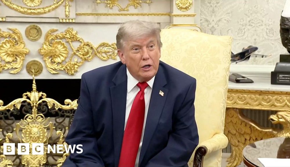 Donald Trump sits in the Oval Office wearing a navy blue suit and red tie.