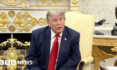 Donald Trump sits in the Oval Office wearing a navy blue suit and red tie.