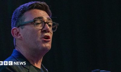 Andy Burnham wearing a black jacket with a black t-shirt talking to an audience with a black background behind him