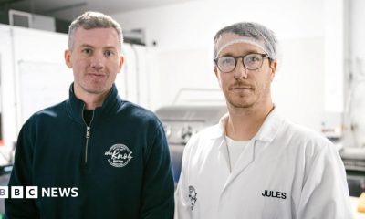 Kieran Woods is wearing a blue fleece with the Knob of Butter logo on it. He is standing next to Jules Bal who is   wearing a hair net, glasses and a white T-shirt with a white overall over it. It has the name Jules sewn into it.