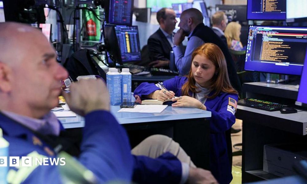 In the left corner, a bald male trader, and a woman with auburn hair, also a trader, in the middle, holding a pen and writing into a notebook, on the floor of the New York Stock Exchange.